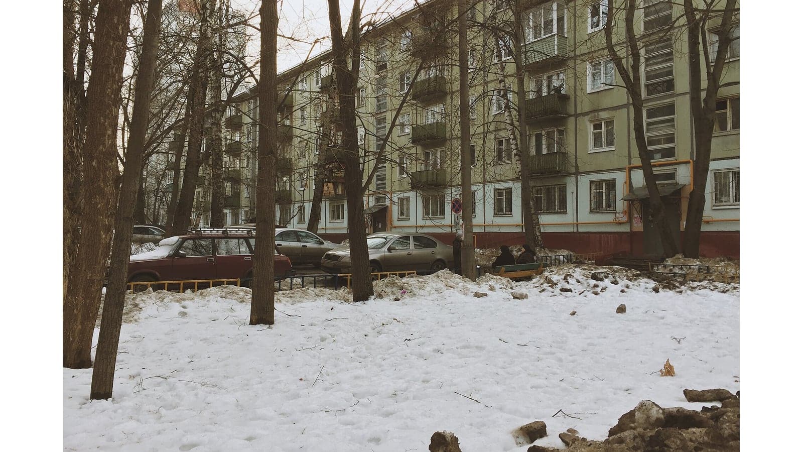 Зимний двор панельной пятиэтажки: деревья, автомобили, скамейка и входы